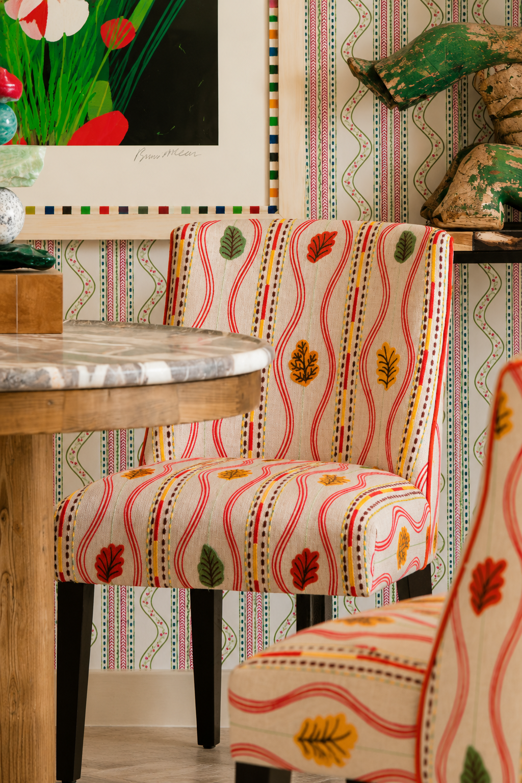 Dining area with upholstered chairs in a colorful Andrew Martin striped botanical fabric, set against coordinating patterned wallpaper beside a marble-topped wood table.