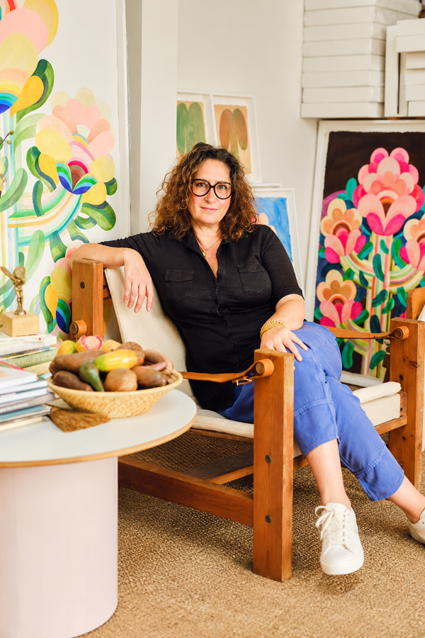 Caroline Rennequin seated in her colorful art studio, surrounded by vibrant floral artwork and paintings in soft pink, green, yellow and blue tones.